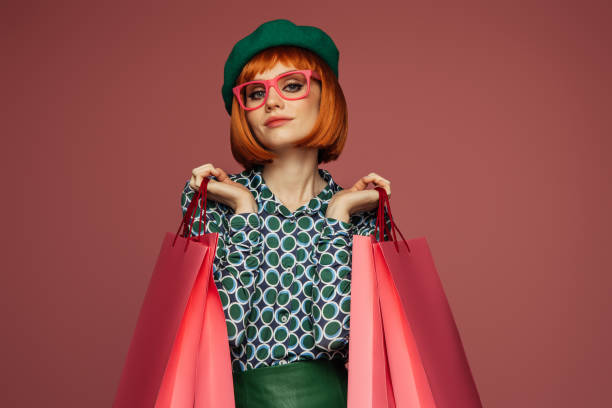 Woman with red hair in stylish outfit holding pink shopping bags against a pink background.